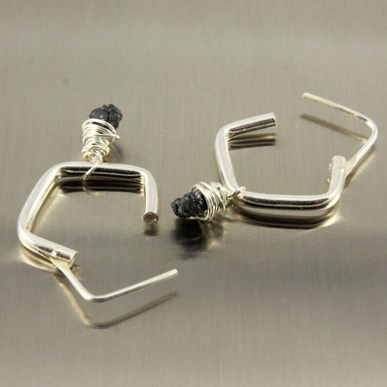 May include: A pair of silver-coloured square hoop earrings. Each earring has a small, dark, rough-textured stone wrapped in silver wire at the top. The hoops have a simple, modern design with a post closure.
