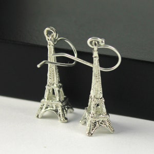 May include: A pair of silver-colored Eiffel Tower earrings. The earrings are detailed replicas of the famous Parisian landmark, featuring intricate architectural details and a hook closure. The earrings are displayed on a white surface.