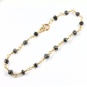 Puede incluir: Una delicada pulsera dorada con piedras preciosas de color gris oscuro y corte rugoso. Las piedras están ensartadas con alambre dorado, creando una joya única y elegante. La pulsera tiene un cierre de broche.