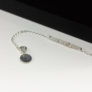 May include: A delicate silver bracelet featuring a strand of small, light gray beads and a small, round charm with the letter "H" engraved on it. The bracelet is displayed on a white surface with a black background.