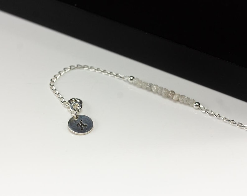 May include: A delicate silver bracelet featuring a strand of small, light gray beads and a small, round charm with the letter "H" engraved on it. The bracelet is displayed on a white surface with a black background.
