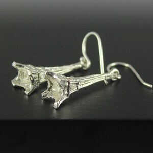 May include: A pair of silver-colored earrings. Each earring features a detailed, stylized trumpet shape with a hook for pierced ears. The earrings are displayed against a dark background, highlighting their metallic sheen.