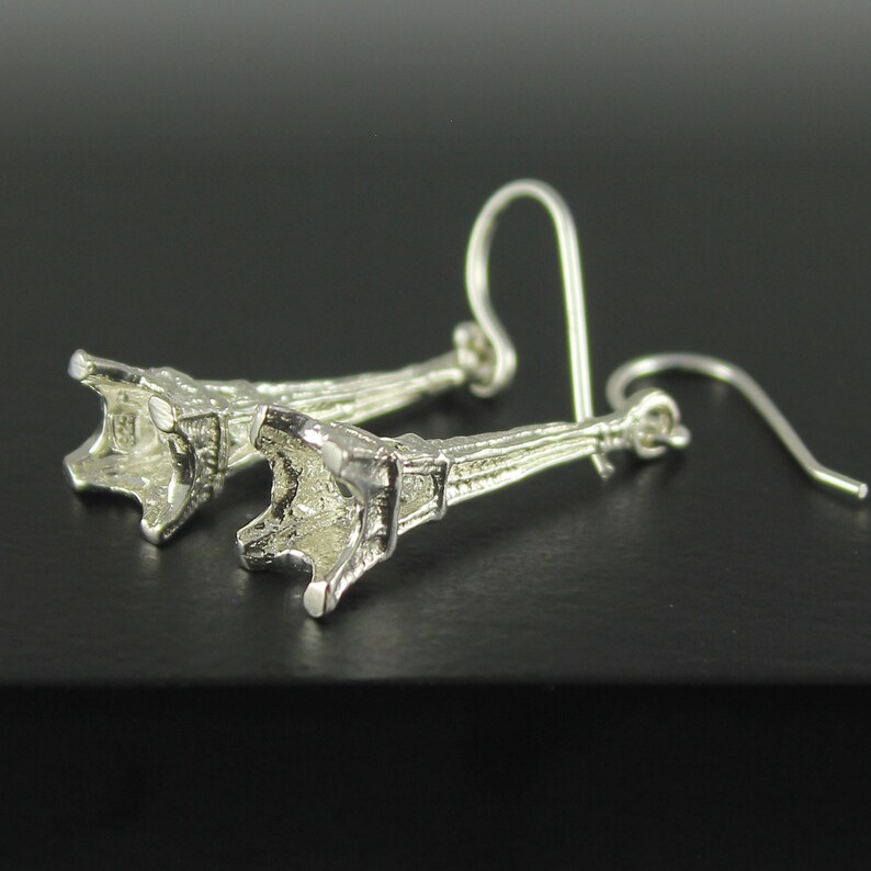 May include: A pair of silver-colored earrings. Each earring features a detailed, stylized trumpet shape with a hook for pierced ears. The earrings are displayed against a dark background, highlighting their metallic sheen.