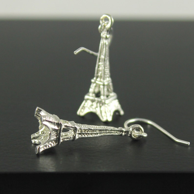 May include: Silver-colored Eiffel Tower earrings. The earrings are detailed miniature replicas of the iconic Parisian landmark. One earring is in focus, showing the hook and the tower's intricate design. The other earring is slightly out of focus in the background.