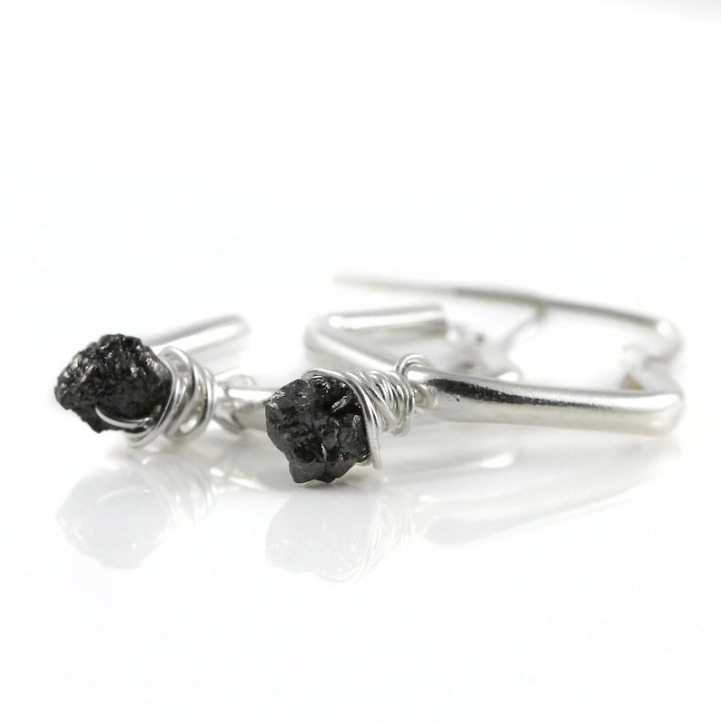 May include: Silver hoop earrings with a raw, black gemstone on each. The gemstones are wrapped with silver wire. The earrings are set against a white background, emphasizing the contrast and the handcrafted nature of the jewellery.