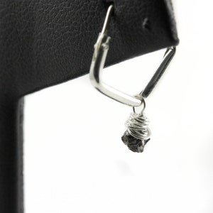 May include: A silver-coloured, geometric earring with a triangular design. A small, dark-coloured stone is wrapped in silver wire and hangs from the earring. The earring is displayed against a white background.