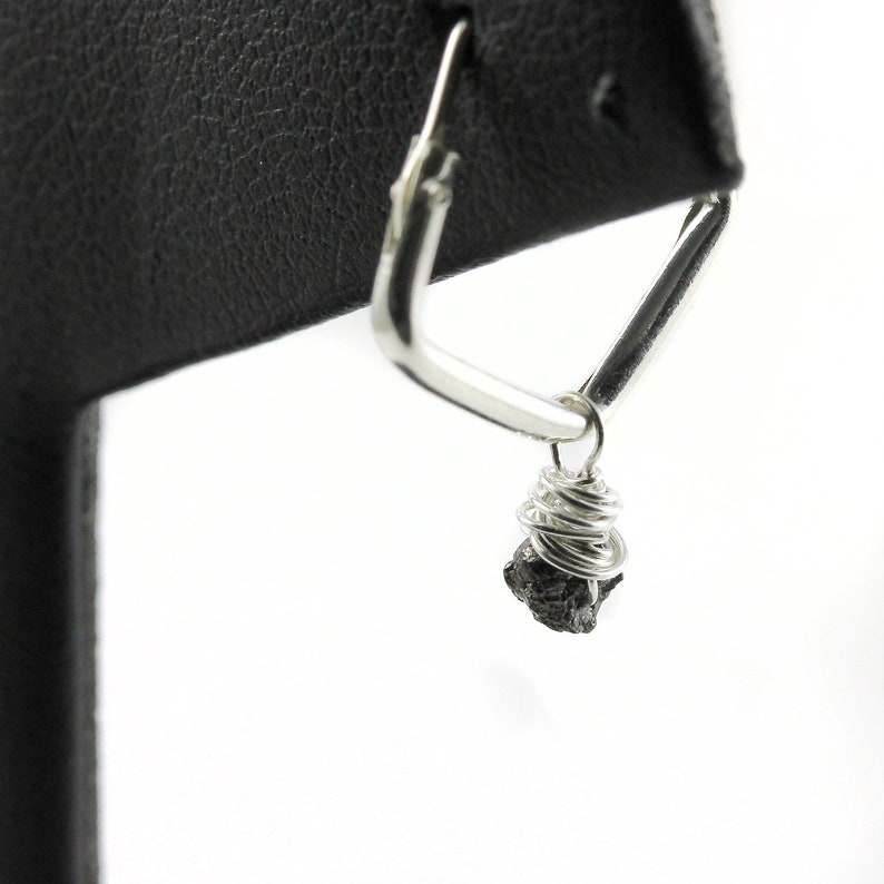 May include: A silver-coloured, geometric earring with a triangular design. A small, dark-coloured stone is wrapped in silver wire and hangs from the earring. The earring is displayed against a white background.