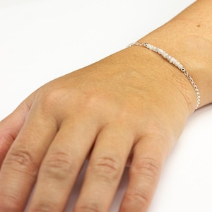 May include: A delicate silver bracelet with small, light-colored beads strung across the center. The bracelet is on a person's wrist, showcasing the jewelry's design and fit. The background is a plain white surface.