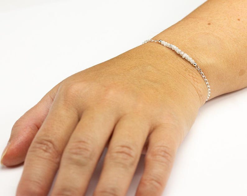 May include: A delicate silver bracelet with small, light-colored beads strung across the center. The bracelet is on a person's wrist, showcasing the jewelry's design and fit. The background is a plain white surface.