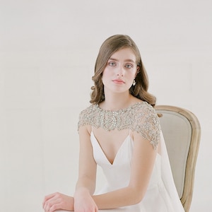 K&ouml;nnte beinhalten: Eine Frau in einem wei&szlig;en Brautkleid mit einem bestickten Spitzen-Schal. Sie sitzt auf einem Stuhl vor einem wei&szlig;en Hintergrund.