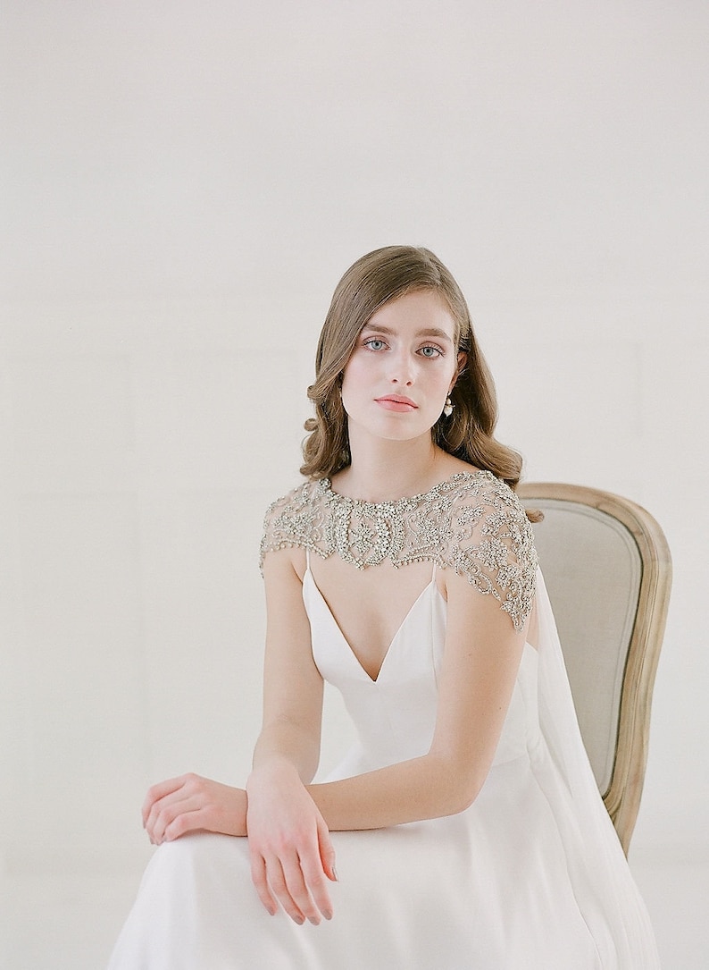 K&ouml;nnte beinhalten: Eine Frau in einem wei&szlig;en Brautkleid mit einem bestickten Spitzen-Schal. Sie sitzt auf einem Stuhl vor einem wei&szlig;en Hintergrund.