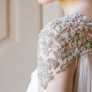 K&ouml;nnte beinhalten: Nahaufnahme eines Brautkleides mit einem perlenbesetzten, floralen Design auf der Schulter. Das Kleid ist aus einem durchsichtigen Stoff gefertigt und hat einen flie&szlig;enden Rock.