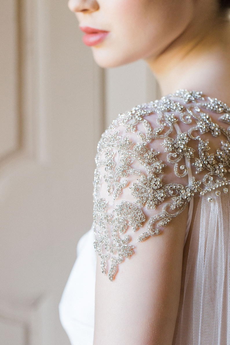 K&ouml;nnte beinhalten: Nahaufnahme eines Brautkleides mit einem perlenbesetzten, floralen Design auf der Schulter. Das Kleid ist aus einem durchsichtigen Stoff gefertigt und hat einen flie&szlig;enden Rock.