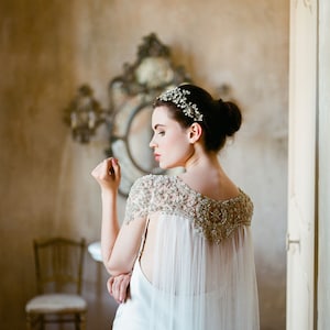 K&ouml;nnte beinhalten: Eine Braut in einem wei&szlig;en Brautkleid mit einer Perlenkappe und einem Juwelenhaarschmuck. Die Braut steht in einem Raum mit einem Spiegel und einem Stuhl.