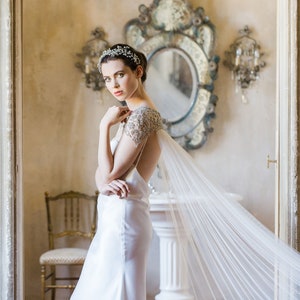 K&ouml;nnte beinhalten: Eine Braut in einem wei&szlig;en Satin-Kleid mit einer bestickten Spitzen-&Uuml;berlage und einem durchsichtigen Schleier. Sie steht in einem Raum mit einem gro&szlig;en, verzierten Spiegel und einem wei&szlig;en Sockel.