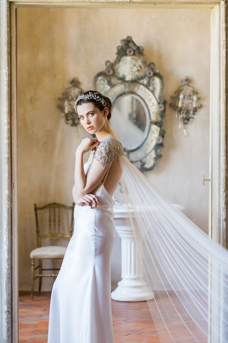 K&ouml;nnte beinhalten: Eine Braut in einem wei&szlig;en Satin-Kleid mit einer bestickten Spitzen-&Uuml;berlage und einem durchsichtigen Schleier. Sie steht in einem Raum mit einem gro&szlig;en, verzierten Spiegel und einem wei&szlig;en Sockel.
