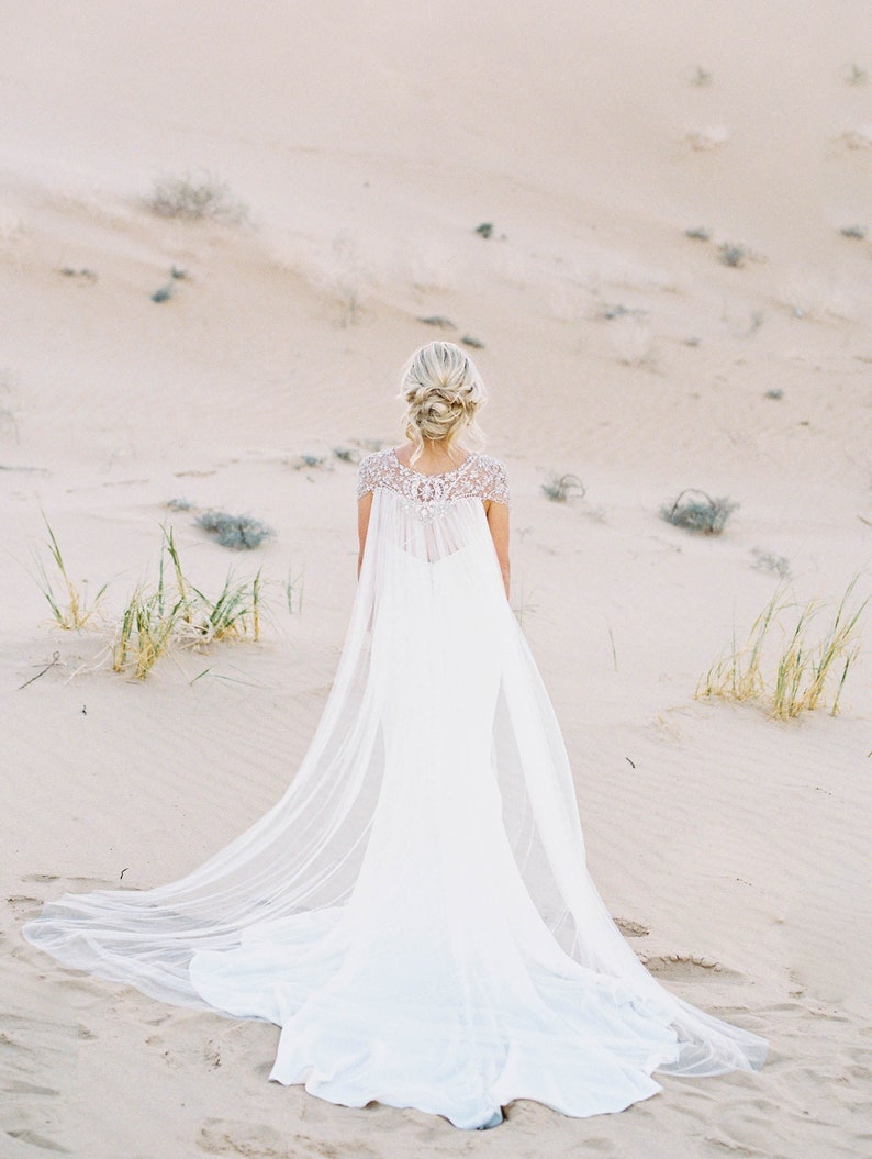 K&ouml;nnte beinhalten: Eine Braut in einem wei&szlig;en Brautkleid mit langem, flie&szlig;endem Schleier und einem perlenbesetzten Oberteil. Das Kleid hat einen durchsichtigen Umhang, der hinter ihr herweht. Sie steht in einer W&uuml;stenlandschaft.