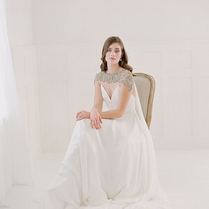 K&ouml;nnte beinhalten: Eine Frau in einem wei&szlig;en Brautkleid mit einer Perlenkappe sitzt auf einem Stuhl. Das Kleid hat einen V-Ausschnitt und einen flie&szlig;enden Rock.