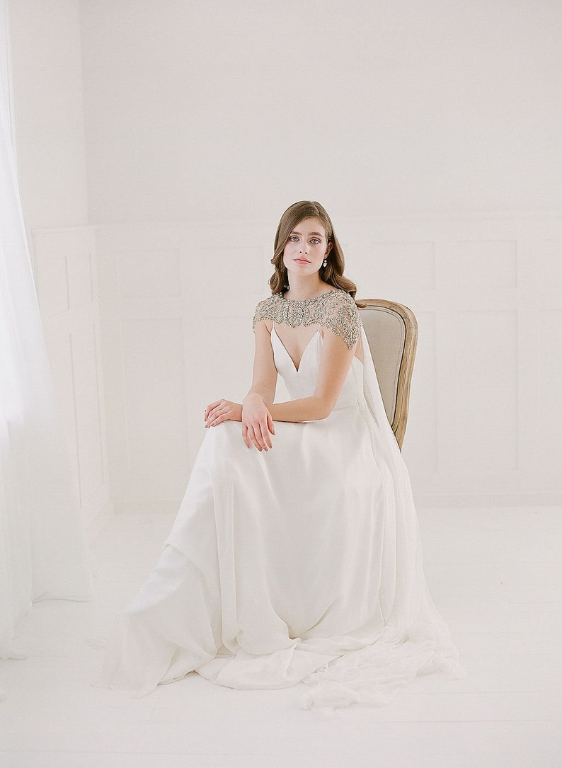 K&ouml;nnte beinhalten: Eine Frau in einem wei&szlig;en Brautkleid mit einer Perlenkappe sitzt auf einem Stuhl. Das Kleid hat einen V-Ausschnitt und einen flie&szlig;enden Rock.