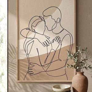 May include: A framed wall art print featuring a black continuous line drawing of two people embracing. The background consists of abstract shapes in shades of beige and tan. The artwork is displayed in a light-colored wooden frame.