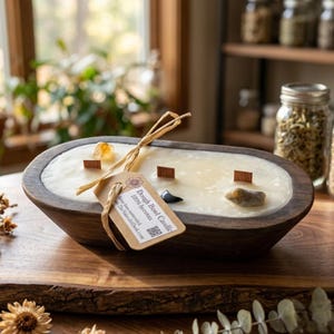 Puede incluir: Un cuenco de madera ovalado de color marrón oscuro lleno de cera de abeja de color blanco roto y tres mechas de madera. La vela incluye cristales incrustados y una etiqueta de papel beige atada con rafia, con el texto Dough Bowl Candle 100% beeswax.