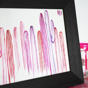 Puede incluir: Una pintura de acuarela enmarcada de una fila de cactus rayados de color rosa, morado y rojo. La pintura est&aacute; sobre un fondo blanco y est&aacute; enmarcada en un marco negro.
