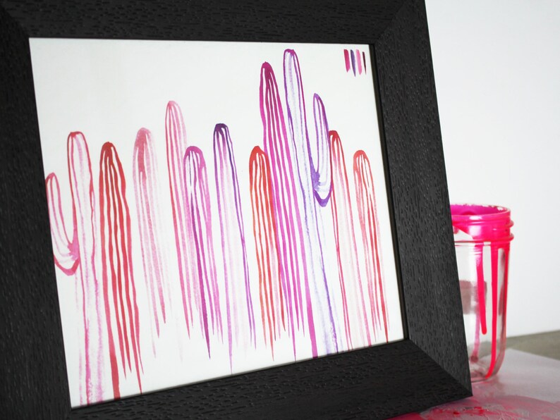 Puede incluir: Una pintura de acuarela enmarcada de una fila de cactus rayados de color rosa, morado y rojo. La pintura est&aacute; sobre un fondo blanco y est&aacute; enmarcada en un marco negro.