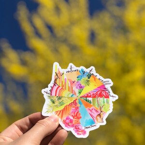 May include: A colorful sticker with a circular design featuring various landscapes and the text "Time spent in nature is time well spent". The sticker is held in a hand against a blurred background of yellow flowers.
