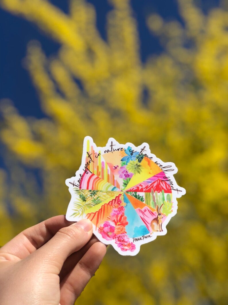 May include: A colorful sticker with a circular design featuring various landscapes and the text "Time spent in nature is time well spent". The sticker is held in a hand against a blurred background of yellow flowers.