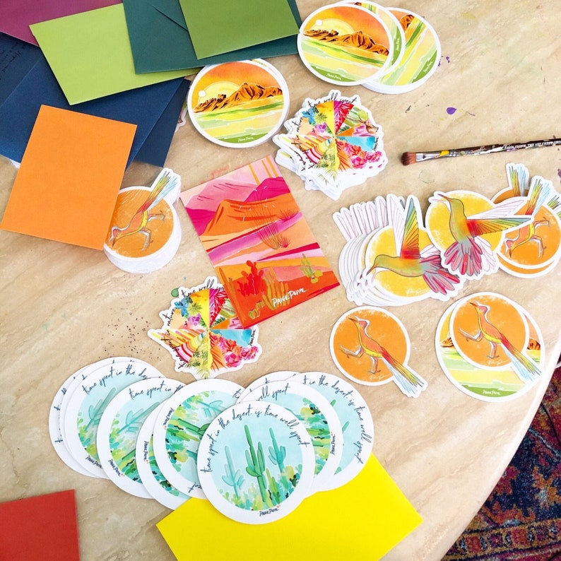 May include: A collection of colorful stickers featuring desert landscapes, birds, and inspirational quotes. The stickers are printed on white paper with a glossy finish. The text on the stickers reads "The best is yet to come" and "Time spent in the desert is time well spent".