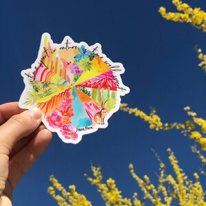 May include: A colorful round sticker with the text "Time spent in nature is time well spent" and a whimsical illustration of a desert landscape with cacti, flowers, and a rabbit. The sticker is held in a hand against a blue sky with yellow branches in the background.