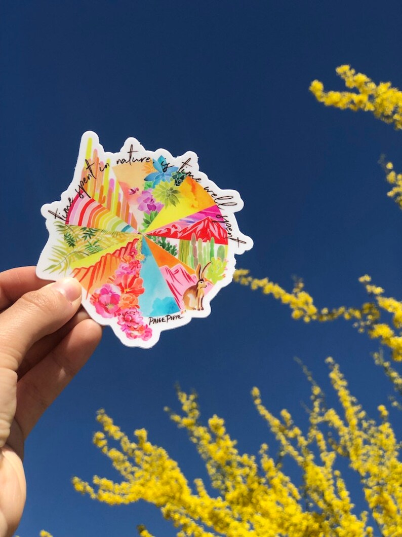 May include: A colorful round sticker with the text "Time spent in nature is time well spent" and a whimsical illustration of a desert landscape with cacti, flowers, and a rabbit. The sticker is held in a hand against a blue sky with yellow branches in the background.