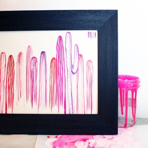 Puede incluir: Una pintura de acuarela enmarcada de una fila de cactus rosas y morados. La pintura est&aacute; en un marco negro y est&aacute; sobre una superficie blanca. Hay un tarro de pintura rosa en el fondo.