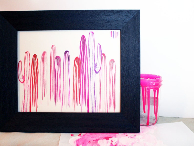 Puede incluir: Una pintura de acuarela enmarcada de una fila de cactus rosas y morados. La pintura est&aacute; en un marco negro y est&aacute; sobre una superficie blanca. Hay un tarro de pintura rosa en el fondo.