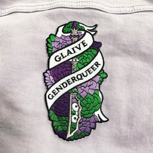 Puede incluir: Un parche bordado con un esquema de color verde, morado y blanco. Presenta una espada estilizada con una pancarta que dice "GLAIVE GENDERQUEER".
