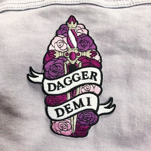 Puede incluir: Parche bordado con un diseño de daga rodeada de rosas moradas y rosas. Una pancarta muestra las palabras "DAGGER DEMI" en negro. El parche está sobre una tela morada clara, adecuado para ropa o accesorios.