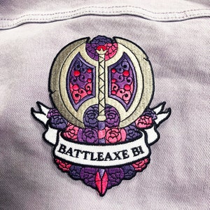 Peut inclure: Un patch brodé doré avec un motif de hache stylisé entouré de fleurs roses et violettes. Le patch porte le texte "BATTLEAXE BI" en blanc.