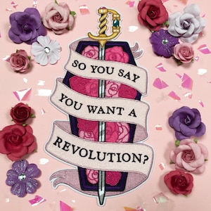 Puede incluir: Un parche bordado rosa y blanco con una espada y un ataúd. El texto en el parche dice "So you say you want a revolution?"
