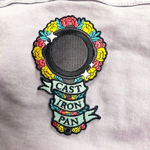 May include: A black and white embroidered patch with a cast iron pan surrounded by a wreath of yellow, red, and blue flowers. The text "CAST IRON PAN" is written on a blue ribbon.