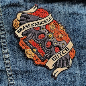 May include: Embroidered patch featuring a brass knuckle design surrounded by roses in shades of red, orange, and yellow. The patch includes a banner with the words "BRASS KNUCKLE BUTCH".