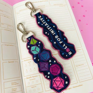 May include: Two embroidered keychains with silver clasps. One reads "ROLL FOR INITIATIVE" in white text on a navy blue background with pink trim. The other features colorful dice shapes. Both are on a notebook page.