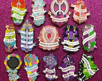 May include: A set of 15 enamel pins featuring various weapons and phrases related to LGBTQ+ identity. The pins are colorful and feature a variety of designs, including a spear, a trident, a shield, a sword, a battleaxe, a glaive, a longbow, a dagger, a polearm, a mace, and a pair of nunchucks. The pins are all about 1 inch in diameter.
