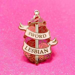 Puede incluir: Pin de esmalte de color dorado con una espada rodeada de rosas rosas y rojas. La espada tiene una hoja blanca y un mango a rayas rojas y blancas. Una pancarta con el texto "SWORD LESBIAN" está envuelta alrededor de la espada y las rosas.