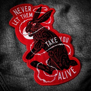 May include: A red and black embroidered patch depicting a rabbit caught in barbed wire. The patch reads "NEVER LET THEM TAKE YOU ALIVE" in white text. The patch is attached to a denim jacket.