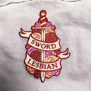 May include: Embroidered patch featuring a sword surrounded by pink and red roses. The text "Sword Lesbian" is on a banner.