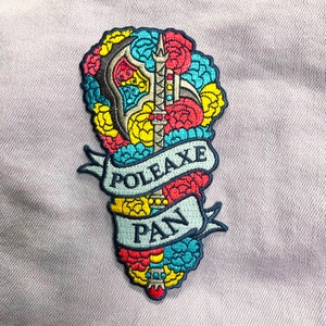 Puede incluir: Un parche bordado colorido con un diseño floral y un arma de asta. El parche tiene las palabras "Poleaxe Pan" bordadas.