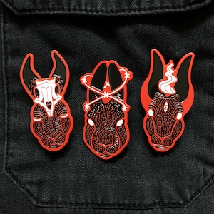 May include: Three black and red enamel pins featuring different rabbit designs. The first pin features a rabbit with a skull and crossbones design. The second pin features a rabbit with a barbed wire design around its head. The third pin features a rabbit with a single eye and a flame design.