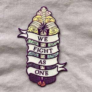 May include: Purple embroidered patch with a sword surrounded by flowers and the text "WE FIGHT AS ONE".