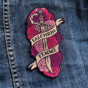 Puede incluir: Parche bordado con una espada plateada con un diseño de rosas rosas y moradas. La espada está rodeada de una pancarta con el texto "Falchion Femme".