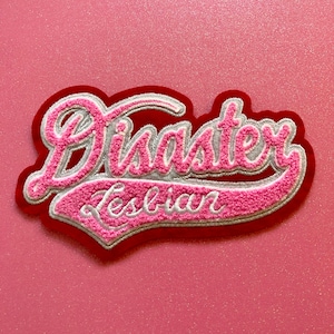 Puede incluir: Parche bordado en rosa y blanco con el texto "Disaster Lesbian" en contorno blanco sobre fondo rojo.